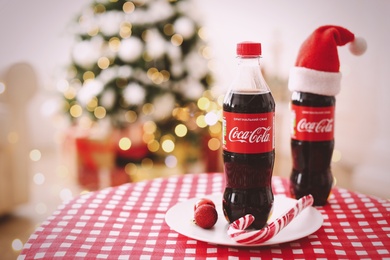 MYKOLAIV, UKRAINE - January 01, 2021: Bottles of Coca-Cola and candy cane on table against blurred Christmas lights