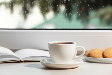 Cup of tea, saucer with cookies and open book on wooden window sill