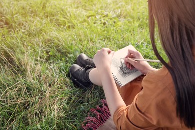 Beautiful young woman drawing with pencil in notepad outdoors on sunny day, closeup