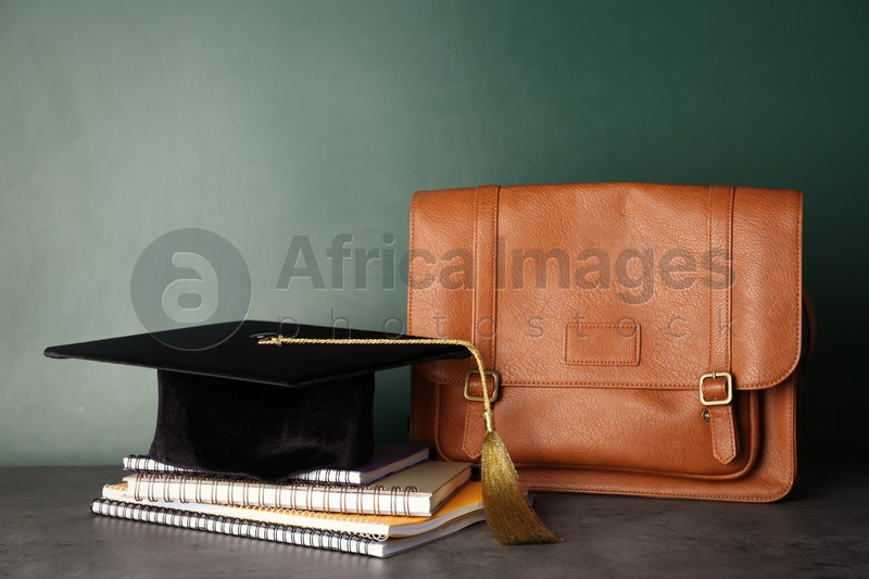 Graduation hat with notebooks and briefcase on table near chalkboard
