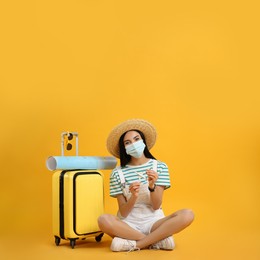 Female tourist in medical mask with suitcase, map and toy plane on yellow background. Travelling during coronavirus pandemic