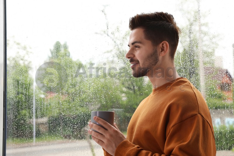 Happy handsome man with cup of coffee near window on rainy day