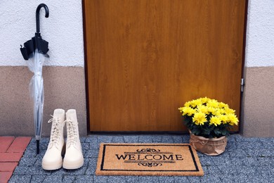 Photo of Doormat with word Welcome, stylish boots, umbrella and beautiful flowers on floor near entrance