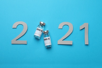 Paper numbers and vials with coronavirus vaccine forming 2021 on light blue background, flat lay
