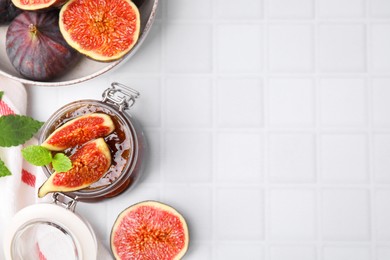 Photo of Glass jar of tasty sweet fig jam and fruits on white tiled table, flat lay. Space for text