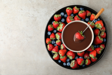 Fondue fork with strawberry in bowl of melted chocolate surrounded by different berries on light table, top view. Space for text