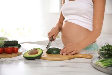 Young pregnant woman cutting avocado at table in kitchen, closeup. Taking care of baby health