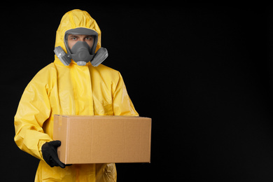 Man wearing chemical protective suit with cardboard box on black background, space for text. Prevention of virus spread