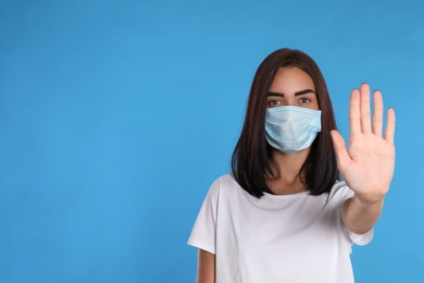 Young woman in protective mask showing stop gesture on light blue background, space for text. Prevent spreading of coronavirus