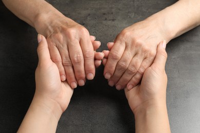 Photo of Young and elderly women holding hands at dark grey table, closeup