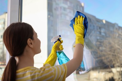 Young woman cleaning window glass with rag and detergent at home, back view