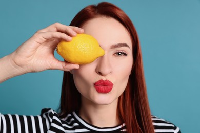 Photo of Beautiful redhead woman covering eye with lemon and blowing kiss on light blue background