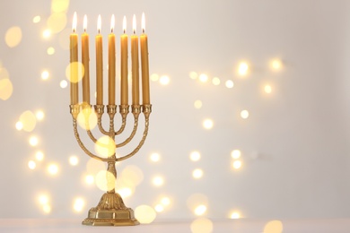 Golden menorah with burning candles against light grey background and blurred festive lights, space for text