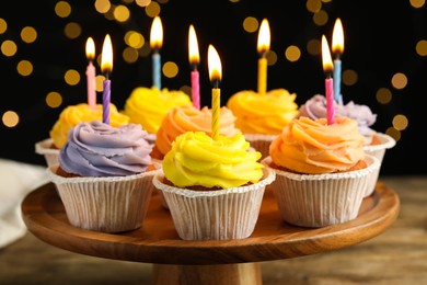 Photo of Tasty birthday cupcakes on wooden stand against blurred lights, closeup