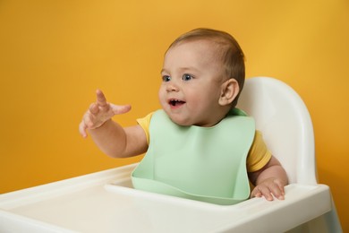 Cute little baby wearing bib in highchair on yellow background