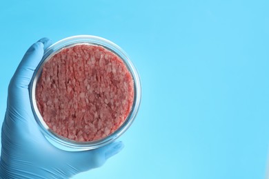 Scientist holding Petri dish with minced lab grown meat on light blue background, closeup. Space for text