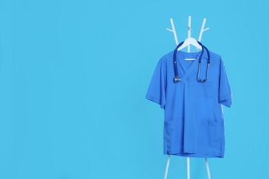 Photo of Medical uniform and stethoscope hanging on rack against light blue background. Space for text