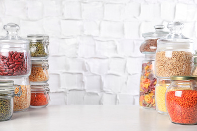 Jars with different cereals on white table against brick wall. Space for text