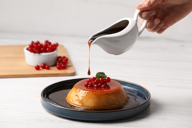 Photo of Woman pouring caramel onto delicious pudding with red currants and mint on white wooden table, closeup