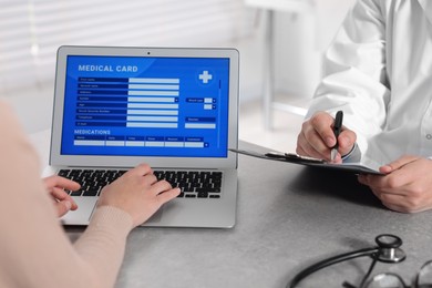 Photo of Doctor and patient using laptop to fill out medical card at table in clinic, closeup