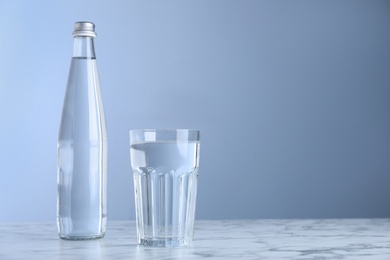Glass and bottle with water on white marble table against blue background, space for text