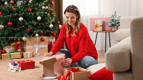 Beautiful young woman wrapping gift near Christmas tree at home