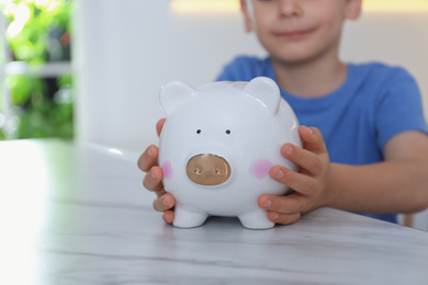 Little boy with piggy bank at marble table indoors, closeup