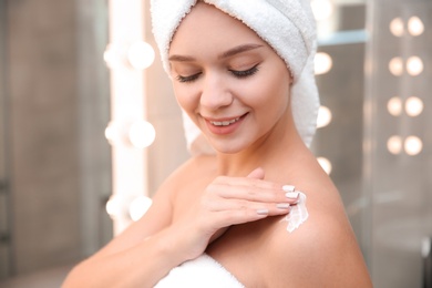 Photo of Beautiful woman with towel on head applying body cream in bathroom