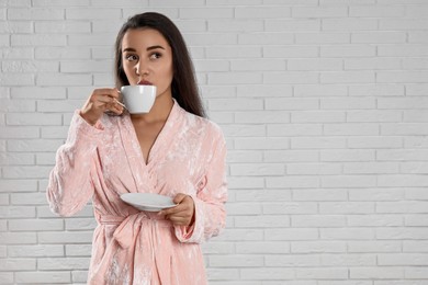 Young woman in bathrobe with cup of coffee near white brick wall. Space for text