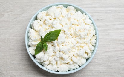 Fresh cottage cheese with basil in bowl on light wooden table, top view