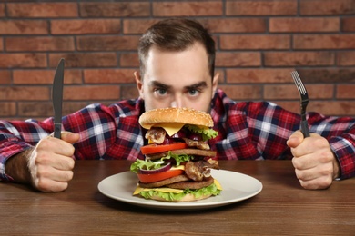 Photo of Young hungry man with cutlery eating huge burger at table
