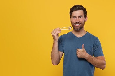 Happy man holding sushi roll with chopsticks and showing thumbs up on orange background. Space for text