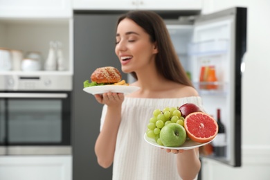 Woman choosing between fruits and burger with French fries near refrigerator in kitchen, focus on food