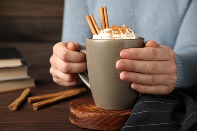 Woman holding cup of hot drink with aromatic cinnamon and whipped cream at wooden table, closeup