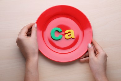 Woman holding plate with paper symbol Ca (Calcium) at wooden table, top view