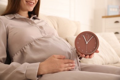 Young pregnant woman holding clock near her belly at home, closeup. Time to give birth