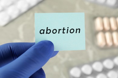 Doctor holding paper card with word ABORTION over table with pills, closeup