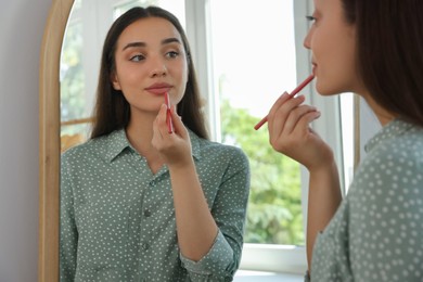 Photo of Beautiful young woman applying cosmetic pencil on lips near mirror indoors