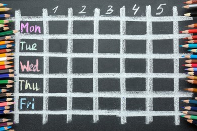 Colorful pencils and drawn weekly school timetable on black chalkboard, top view