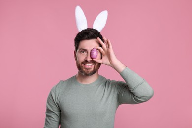 Photo of Happy man in cute bunny ears headband covering eye with Easter egg on pink background