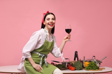 Young housewife with glass of wine, vegetables and different utensils on pink background