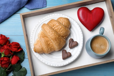 Romantic breakfast with cup of coffee, croissant and roses on blue wooden table, flat lay. Valentine's day celebration