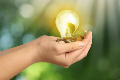 Solar energy concept. Woman holding glowing light bulb with seedling and coins against green blurred background, closeup