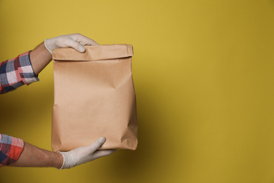 Courier in protective gloves holding paper bag on yellow background, closeup with space for text. Food delivery service during coronavirus quarantine