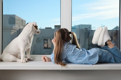 Photo of Cute little girl with her dog on window sill indoors. Childhood pet