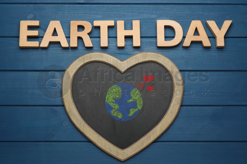 Words Earth Day and heart shaped blackboard with planet drawing on blue wooden table, flat lay