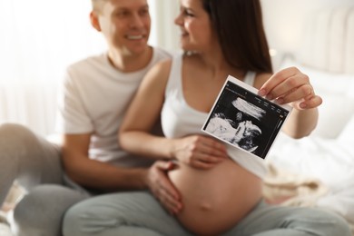 Photo of Young pregnant woman with her husband in bedroom, focus on ultrasound picture of baby
