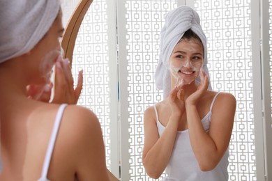 Photo of Beautiful teenage girl applying cleansing foam onto face near mirror at home. Skin care cosmetic