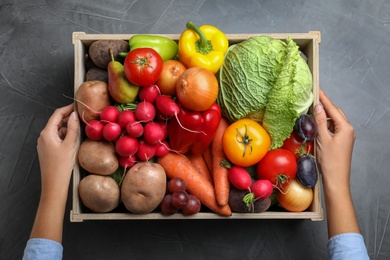 Farmer with wooden crate full of different vegetables and fruits at grey table, top view. Harvesting time