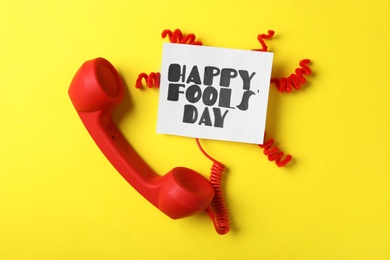Telephone handset and Happy Fools' Day note on yellow background, flat lay